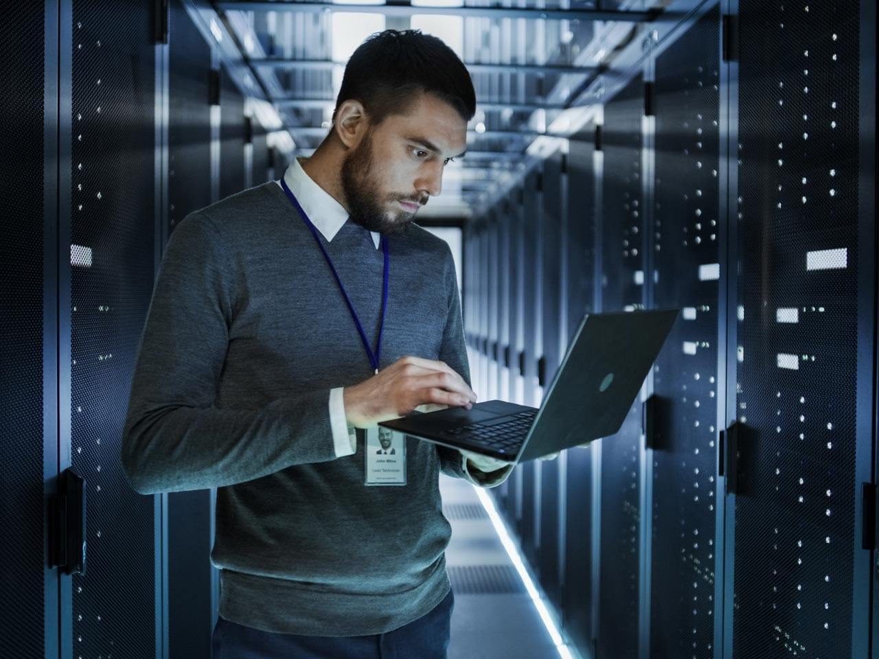 An IT technician standing in a modern data centre corridor, working on a laptop while surrounded by server racks